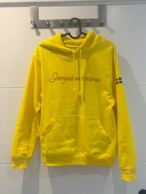 Gul hoodie med Sverigedemokraterna-tryck - Gul hoodie med huva och känguruficka framtill. Trycket 'Sverigedemokraterna' syns på bröstet och en liten svensk flagga finns på ena ärmen. Klassisk passform och ribbade muddar. 