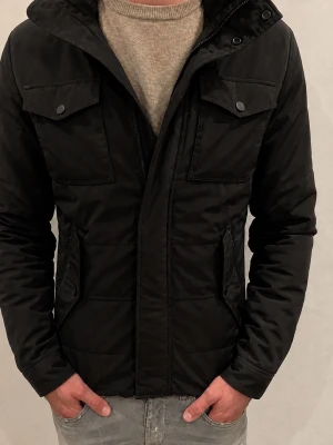 J.Lindeberg Svart Field Jacka - Svart J.Lindeberg Field Jacket i storlek M. Jackan är vadderad och har huva, två bröstfickor samt två sidofickor. Observera att jackan säljs med tydliga defekter: två-tre knappar framtill saknas och dragkedjan fungerar inte, vilket innebär att jackan inte går att stänga framtill i nuvarande skick. Säljs därför som defekt och till ett lägre pris. I övrigt ser jag inga andra större fel. Se bilderna noggrant före köp och skriv gärna vid frågor.