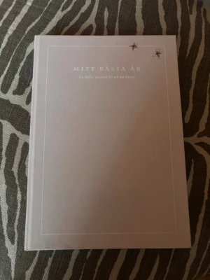 Mitt bästa år - Bullet journal - En stilren bullet journal med titeln 'Mitt bästa år'. Omslaget är ljusrosa med minimalistisk design och små illustrationer. Perfekt för dig som vill planera och reflektera över ditt år på ett kreativt sätt. Innehåller inspirerande sidor för mål och välmående.
