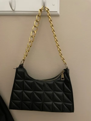 Svart handväska med guldkedja - Svart handväska med en chunky guldfärgad kedja som axelrem. Väskan har dragkedja upptill och en stilren, rektangulär form. Perfekt accessoar för att lyfta din outfit.