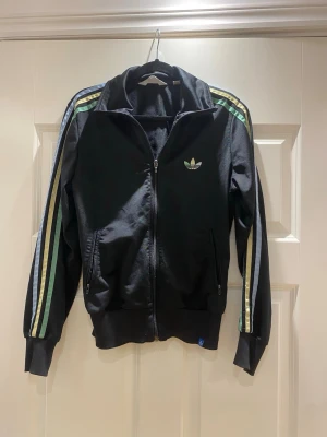 Svart Adidas hoodie med ränder - Snygg svart hoodie från Adidas med dragkedja framtill och klassiska tre ränder i grönt, blått och gult längs ärmarna. Adidas-logga på bröstet och två fickor med dragkedja. Perfekt för en sportig och avslappnad stil.