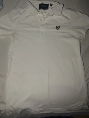 Vit pikétröja från Lyle & Scott - Klassisk vit pikétröja från Lyle & Scott med gul logga på bröstet och krage med två knappar. Tillverkad i bomull och har korta ärmar. Perfekt för en clean och stilren look.