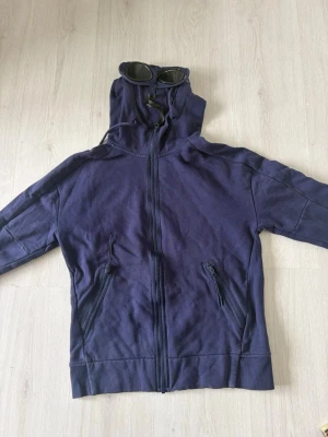 Mörkblå hoodie med glasögon i huvan - Säljer en mörkblå hoodie från C.P. Company med dragkedja framtill och två snedställda fickor med dragkedja. Huvan har inbyggda glasögon och dragsko för justering. Perfekt för dig som vill sticka ut med en unik streetwear-look.