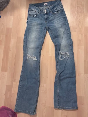 Blå bootcut jeans med slitningar - Säljer ett par ljusblå bootcut jeans med slitna detaljer på båda knäna. Jeansen har klassisk femficksdesign, knappgylf och bakfickor knapp. Slitna vid nederkant
