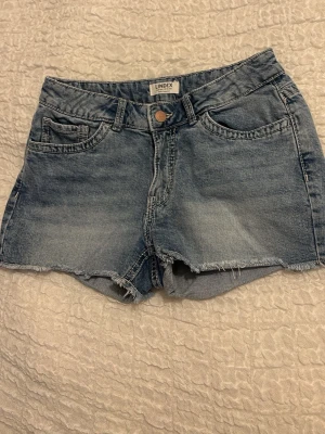 Blå jeansshorts från Lindex - Snygga blå jeansshorts från Lindex med rå kant nertill och klassisk femficksdesign. Shortsen har knapp och dragkedja framtill samt bälteshällor. Perfekta för varma dagar och enkel att matcha med olika toppar.