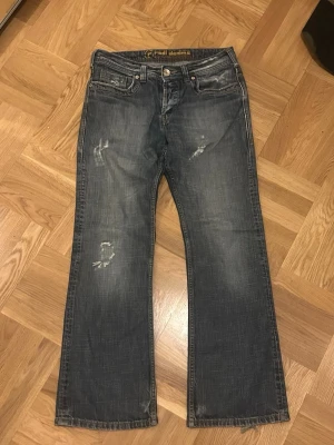 Blå slitna bootcut jeans - Ett par blå bootcut jeans med slitningar på låren och knäna. Jeansen har fem fickor, knappgylf och klassisk denimlook. Materialet är denim och passformen är något utsvängd nedtill. Perfekta för en avslappnad och trendig stil.