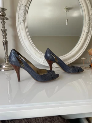 Mörkblå Nine West slingback pumps I läder - Snygga mörkblå pumps från Nine West med slingback och öppen tå. Skorna har krokodilpräglat mönster och bruna klackar. Justerbart spänne på sidan och guldfärgad innersula ger en elegant touch. Perfekta för dig som vill sticka ut.