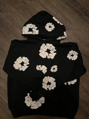 Denim Tears Hoodie - Säljer en svart hoodie med stora vita blommönster över hela plagget, inklusive huvan. Hoodien har en avslappnad passform och är tillverkad i mjukt material, perfekt för en chill och trendig look. Huvan ger extra stilpoäng och värme under kyliga dagar.
