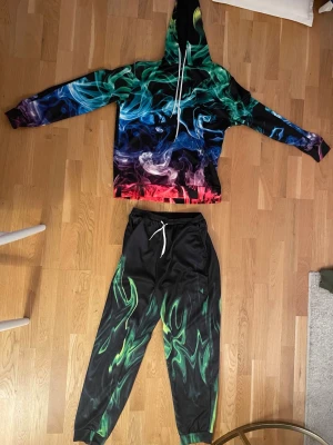 Färgstarkt loungewear set - Säljer ett loungewear set med hoodie och byxor i mjukt material. Setet har ett unikt rök- och eldliknande tryck i grönt, blått, rött och svart. Hoodien har dragsnören och byxorna har elastisk midja med vita snören. Perfekt för chill dagar hemma eller när du vill sticka ut. Storlek är S 