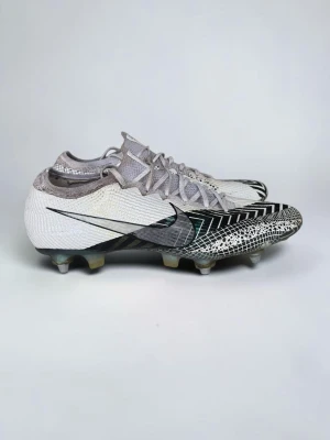 Vita Nike Mercurial fotbollsskor - Säljer ett par vita Nike Mercurial fotbollsskor med svart sicksackmönster och grafiska detaljer. Skorna har snörning, låg profil och är tillverkade i ett lätt syntetmaterial för bästa bollkontroll och grepp på planen. Perfekta för dig som vill sticka ut på matchen.