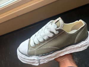Maison Mihara Yasuhiro MMY - Size 42 43. Unika sneakers i olivgrönt tyg med breda vita skosnören och en chunky, vågig vit sula. Skon har vit tåhätta i gummi och detaljerade sömmar längs sidan. Perfekt för dig som vill sticka ut med din stil. Helt nya 