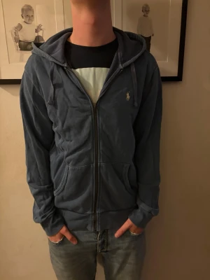 Blå zip hoodie med huva - Säljer en blå hoodie med dragkedja och huva. Hoodien har fickor framtill och en liten broderad logga på bröstet. Mjuk och skön, perfekt till chill dagar. Passar till jeans eller joggers.