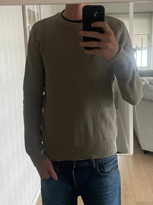 Beige stickad tröja med rund hals - En stilren beige stickad tröja med rund hals och långa ärmar. Tröjan har en enkel, ribbad struktur och sitter snyggt på kroppen. Perfekt att matcha med jeans för en avslappnad look.