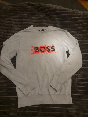 HUGO BOSS TRÖJA - OBS! Ingen fläck på koftan det är min kamera som är trasig!🙌 Snygg grå sweatshirt från HUGO BOSS med ett stort svart och rött tryck på bröstet. Tröjan har rund halsringning, långa ärmar och ribbade muddar vid ärmslut och nederkant. Perfekt för en avslappnad och trendig stil. 