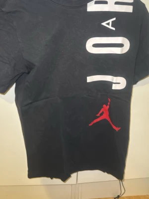 Svart Jordan t-shirt med tryck - Snygg svart t-shirt med stort vitt Jordan-tryck längs framsidan och baksidan samt den klassiska röda Jumpman-loggan. T-shirten har korta ärmar och är gjord i mjuk bomull, perfekt för en sportig och avslappnad stil.