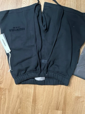 Svarta mjukis shorts  från Essentials - Svarta mjukisbyxor från Essentials med dragsko i midjan och elastiska benslut. Byxorna har en stilren design med diskret logga på benet och är tillverkade i mjukt bomullsmaterial. Perfekta för en avslappnad och trendig look.