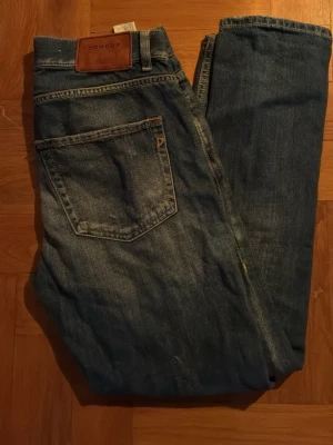 Blå slitna jeans från Dondup - Säljer ett par blå jeans Mila från Dondup  med raka ben och slitningar på båda knäna. Jeansen har klassisk femficksdesign och en läderpatch med logga bak i midjan. Perfekta för en avslappnad och trendig look.