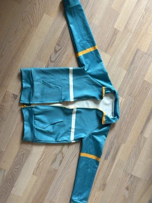 Max Mayfield jacket - Snygg turkos hoodie med dragkedja, vita ränder över bröst och rygg samt gula detaljer på ärmar och vid dragkedjan. Två fickor framtill och mjukt innerfoder. Perfekt för en avslappnad och sportig stil.