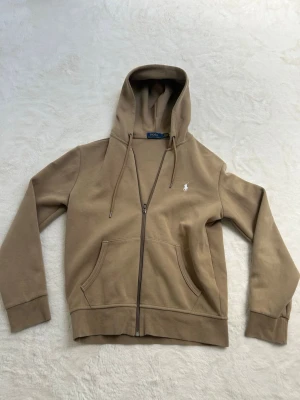 Beige hoodie med dragkedja från Polo Ralph Lauren - Snygg beige hoodie från Polo Ralph Lauren med dragkedja framtill och klassisk huva. Har två fickor fram och den ikoniska lilla loggan broderad på bröstet. Perfekt för en chill och stilren look.