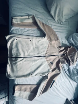 Beige och vit hoodie från Nike man få byxorna med  - Snygg hoodie från Nike i beige och vitt med dragkedja framtill och två fickor. Hoodien har en huva och diskreta svarta Nike-loggor på bröstet och ärmen. Tillverkad i mjukt material som känns skönt mot huden. Perfekt för en avslappnad och sportig stil.