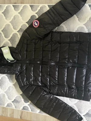 Svart dunjacka från Canada Goose - Snygg svart dunjacka från Canada Goose med huva och klassisk logotyp på ärmen. Jackan har quiltad design, dragkedja framtill och två sidofickor med dragkedja. Perfekt för kalla vinterdagar och riktigt lätt att bära.
