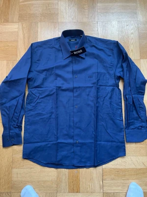 Marinblå skjorta från Hugo Boss - Snygg marinblå skjorta från Hugo Boss med klassisk krage och diskret logga på bröstet. Skjortan har långa ärmar och knäppning framtill. Perfekt för dig som vill ha en stilren och tidlös look.