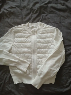 Vit quiltad cardiganjacka - Snygg vit cardiganjacka med quiltad framsida och stickade ärmar. Jackan har dragkedja framtill, ribbade muddar och två fickor på magen. Perfekt för lager på lager och ger en clean look till din outfit.