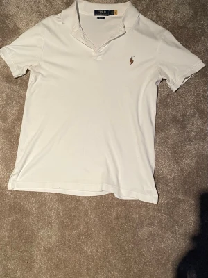 Vit pikétröja från Polo Ralph Lauren - Vit pikétröja från Polo Ralph Lauren i slim fit-modell. Klassisk krage, korta ärmar och broderad logga på bröstet. Perfekt till jeans eller shorts för en clean och stilren look.