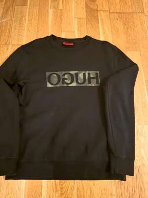 Svart sweatshirt med HUGO-logga - Svart sweatshirt med rund hals och lång ärm från HUGO. På bröstet finns en stor logga i svart text mot en camouflagemönstrad bakgrund. Tröjan har ribbade muddar vid ärmslut och nederkant. Perfekt för en stilren och trendig look.