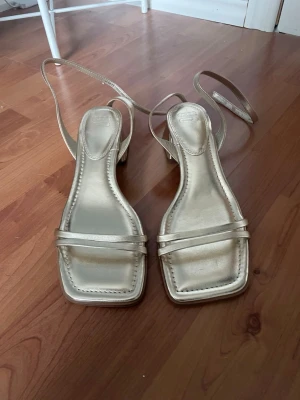 Guldiga sandaletter med fyrkantig tå - Snygga sandaletter i metallic guld med smala remmar och fyrkantig tå. Skorna har en låg, bred klack och långa remmar som kan snöras runt ankeln. Tillverkade i ett glansigt syntetmaterial som ger en trendig look.