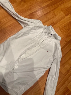 Vit skjorta från Tommy Hilfiger - Den är 140 cm lång Klassisk vit skjorta från Tommy Hilfiger med liten broderad logga på bröstet. Skjortan har lång ärm, knappar framtill och är tillverkad i bomull. Perfekt till många olika stilar och enkel att matcha.