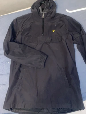 vindjacka från lyle scout  - Snygg svart vindjacka med huva och halv dragkedja framtill. Jackan har en gul logga på bröstet, stor magficka och elastiska muddar vid ärmsluten. Perfekt för blåsiga dagar och enkel att matcha med olika outfits.