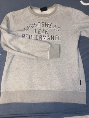 Grå sweatshirt Peak Performance - Säljer en ljusgrå sweatshirt från Peak Performance med texten 'SPORTSWEAR PEAK PERFORMANCE' broderad framtill. Tröjan har rund halsringning, långa ärmar och ribbade muddar vid ärmslut och nederkant. Perfekt för en avslappnad stil.