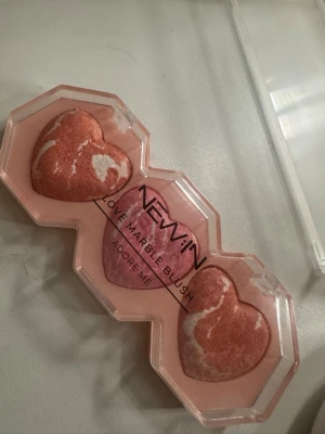 Hjärtformat marmorerat rouge - Rougepalett med tre hjärtformade marmorerade rouge i rosa och persikotoner. Kommer i en transparent plastförpackning med texten 'NEW'IN LOVE MARBLE BLUSH ADORE ME' på locket. Perfekt för att ge kinderna en fräsch glow och färg.