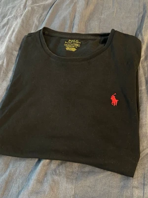 Svart t-shirt från Polo Ralph Lauren - Säljer en svart t-shirt från Polo Ralph Lauren med röd broderad logga på bröstet. Modellen är custom slim fit och har rund halsringning. Tillverkad i mjuk bomull som känns skön mot huden. Perfekt för dig som gillar klassisk och stilren design.OBS, den är äkta!