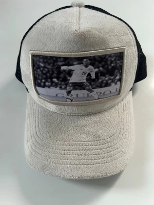 Vit och svart keps med fotomotiv - Säljer en stilren keps från Evosportswear i vitt och svart med mesh på baksidan. Framsidan har ett svartvitt fotomotiv av en fotbollsspelare. Kepsen har böjd skärm och justerbar passform. Perfekt för dig som gillar sportig stil.