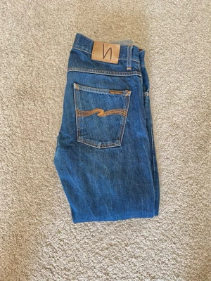 Nudie Jeans | Grim Tim - Dry orange selvedge - Säljer ett par klassiska blå Grim Tims  från Nudie Jeans med snygga kontrastsömmar och ikonisk bakficka med vågig detalj. Byxorna är selvedge vilket medger att användandet av byxorna gör dom till ett unikt par jeans varje gång. Byxorna har blivit sydda( se bild två). Det är 32/32 men pågrund av tvätt och annat så skulle jag säga 31/32, 30,5/32