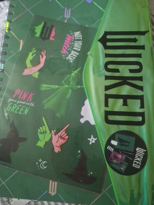 Wicked anteckningsbok med klistermärken - Säljer en spiralbunden anteckningsbok med grönt omslag och motiv från musikalen Wicked. Medföljer klistermärken med häxhattar, händer och citat som 'Not your basic witch' och 'Pink goes good with green'. Perfekt för fans av musikaler och fantasy.