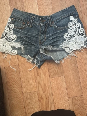 Jeansshorts med spetsdetaljer - Säljer ett par blå vintage jeansshorts med råa kanter och vita spetsapplikationer på framsidan. Shortsen har fem fickor, bälteshällor och dekorativa sömmar på bakfickorna. Perfekta för sommaren och ger en boho-vibe.