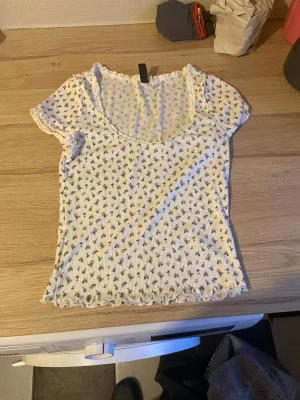 Vit blommig ribbad topp - Söt vit ribbad topp med små svarta blommor och korta ärmar. Toppen har en rundad halsringning och vågig kant nertill och vid ärmslut. Perfekt för en trendig och avslappnad look.