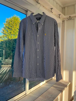 Randig skjorta från Polo Ralph Lauren - Klassisk blåvit randig skjorta från Polo Ralph Lauren med gul broderad logga på bröstet. Skjortan har lång ärm, knappar framtill och krage. Perfekt till jeans eller chinos för en clean och stilren look.