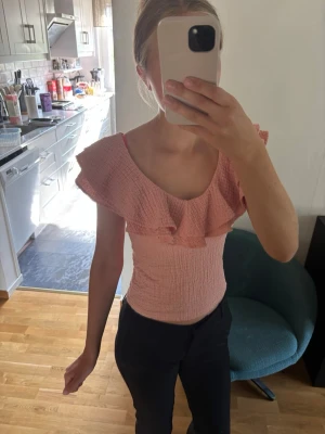 Ljusrosa volangtopp - Supersöt ljusrosa topp med smockad passform och bred volang runt axlarna. Toppen är ärmlös och har en figurnära, stretchig modell som sitter snyggt på kroppen. Använd 2-3 gånger och är precis som ny! Säljer för att den är för liten för mig.