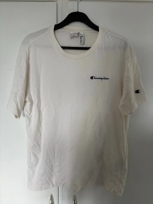Vit Champion t-shirt med logga - Vit t-shirt från Champion med klassisk logga broderad på bröstet och ärmen. T-shirten har rund hals och korta ärmar. Tillverkad i mjuk bomull för en skön och avslappnad stil. Bra skick 