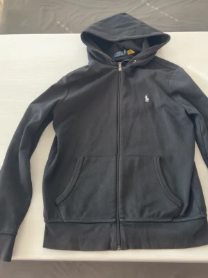 Svart hoodie med dragkedja från Polo - Snygg svart hoodie från Polo Ralph Lauren med dragkedja framtill och klassisk vit logga på bröstet. Hoodien har två fickor fram, ribbade muddar och en skön huva. Tillverkad i mjukt bomullsmaterial som passar perfekt till en avslappnad stil.
