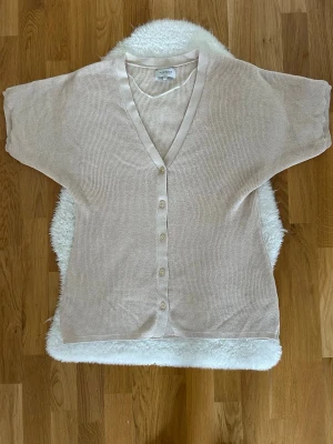 Beige kofta från Alice Bizous - Ljusbeige kofta från Alice Bizous med korta ärmar och v-ringning. Koftan har knappar framtill och är tillverkad i ett tunt, stickat material som känns luftigt och skönt. Perfekt att slänga över en topp för en avslappnad look.
