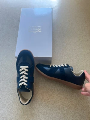 Maison Margiela gats - Säljer ett par mörkblå sneakers från Maison Margiela med vita snören och brun sula. Skorna är helt oanvända och är storlek 43. Skriv vid frågor och fler bilder, pris kan diskuteras vid snabb affär