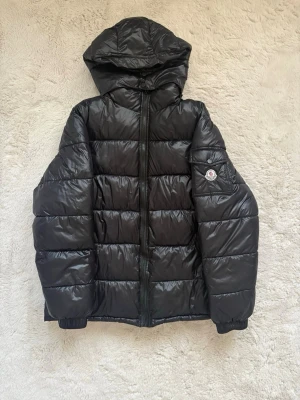 Svart Moncler jacka - Snygg svart dunjacka från Moncler med glansig finish, huva och dragkedja framtill. Jackan har två fickor med dragkedja och en Moncler-logga på ärmen. Pris kan diskuteras.
