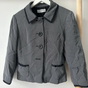 Grå kavaj med svarta detaljer - Stilren grå kavaj med diskret mönster och svarta kontrastdetaljer vid krage, fickor och knappslå. Tre stora knappar framtill och två fickor. Saknar en fjärde knapp. Övrigt i väldigt bra skick. USA storlek 8, motsvarar storlek M.