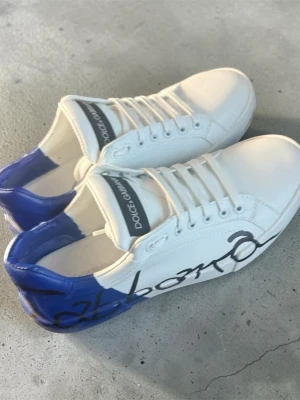  vita sneakers blå detalj - Snygga vita sneakers med blå häl och svart grafisk text på sidan. Skorna har vita snören, logga på plösen och sula med präglad märkestext. Tillverkade i läder för en exklusiv känsla. Använda en gång. Hör av er!!