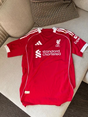 Liverpool FC hemmatröja – Adidas - Röd Liverpool FC fotbollströja från Adidas med Standard Chartered-sponsor och Expedia på ärmen. Klassisk röd/vit design med klubbmärke och Adidas-detaljer. Authentic Licensed Product-märkning nedtill.  Fint skick utan tydliga större skador eller fläckar. Storlek syns inte på bilderna, så skicka gärna bild på storleksetiketten om du vill att jag lägger in den också.